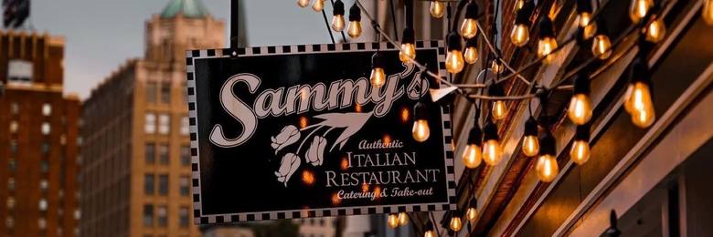 Sammy's hanging sign over 3rd Street at dusk, strung with warm lights